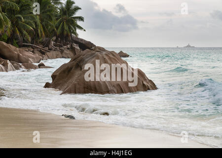 Schöner Strand Anse Patates, La Digue, Seychellen mit Granitfelsen an einem bewölkten Tag, einer kleinen Insel im Hintergrund Stockfoto