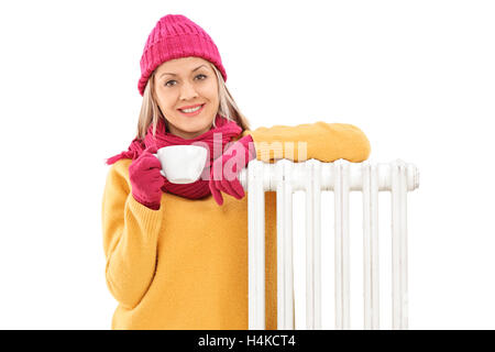 Fröhliche Frau, stützte sich auf einen Heizkörper und hält eine Tasse isoliert auf weißem Hintergrund Stockfoto