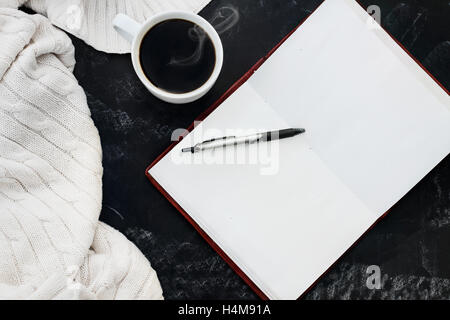 Weiche Strickpullover Decke mit einer heißen Tasse Kaffee und ein offenes Buch mit Stift über Grunge Tafel Hintergrund. Stockfoto