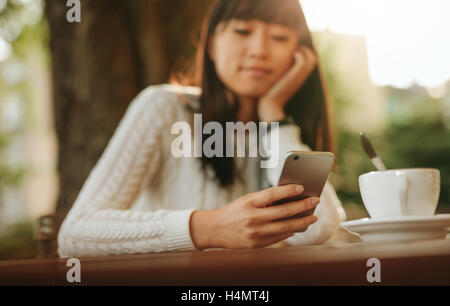 Junge Chinesin Blick auf Handy in der Hand beim Sitzen im Café im Freien. Asiatische weibliche Modell lesen Text Nachricht o Stockfoto