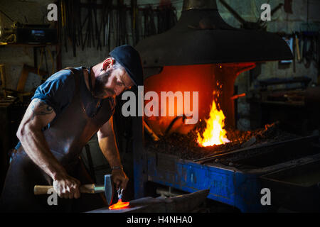 Ein Schmied schlägt ein Stück rote Roheisen am Amboss mit einem Hammer in eine Werkstatt. Stockfoto