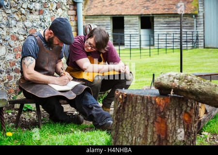 Zwei Schmiede, ein Mann und eine Frau tragen Schürzen in ein Notizbuch schreiben saß in einem Garten. Stockfoto