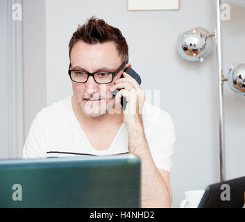 Ein Mann sitzt an einem Schreibtisch im Büro vor einem Laptop, auf dem Handy. Stockfoto