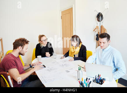 Vier Menschen bei einem Geschäftstreffen an einem Tisch zu verbreiten mit Papieren, ein Mann mit einem Tablett und einer auf einem Laptop. Stockfoto