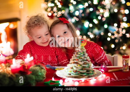 Kleine Jungen und Mädchen machen Weihnachten Lebkuchenhaus am Kamin in eingerichteten Wohnzimmer. Kinder spielen mit Lebkuchen Stockfoto