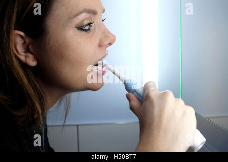 Junge Frau in Badezimmerspiegel zu schauen, wie sie ihre Zähne mit der elektrischen Zahnbürste reinigt Stockfoto