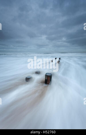 Holzpfosten von einem alten Buhne am Strand als die Nordsee rauscht in bei Flut an der Northumberland Küste an einem grauen Tag Stockfoto