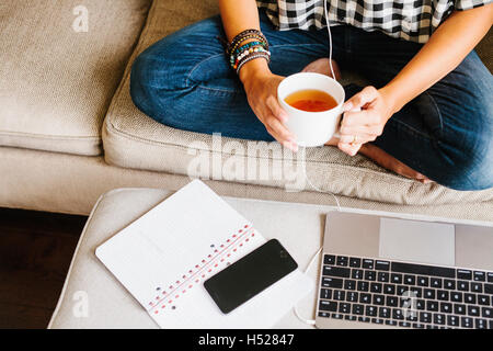 Frau trägt Jeans sitzen auf einem Sofa vor einem Laptop-Computer hält einen Becher. Stockfoto