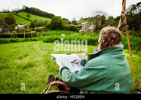 Eine Künstlerin, die mit Kohle auf Papier, sitzen auf dem Boden im Freien skizzieren. Stockfoto