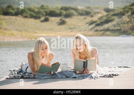 Zwei blonde Schwestern liegen auf einem Steg, ein Buch zu lesen. Stockfoto