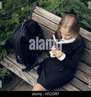 Schöne blondes Mädchen in Schuluniform sitzt auf der Parkbank und Lesebuch. Stockfoto