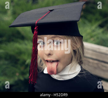 Schöne blondes Mädchen in Schule Uniform und Graduierung Kappe auf dem Kopf ragt Zunge als Albert Einstein. Stockfoto