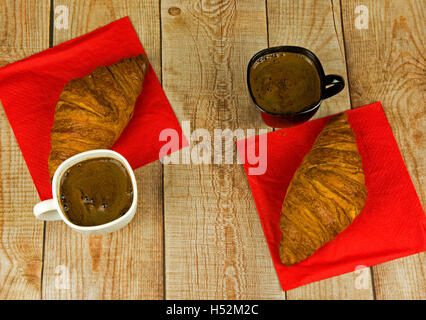 Zwei Croissants und zwei Tassen Kaffee auf rotem Papierservietten, stehen auf einem Holztisch mit Desek. Flache horizontale Ansicht von oben. Stockfoto