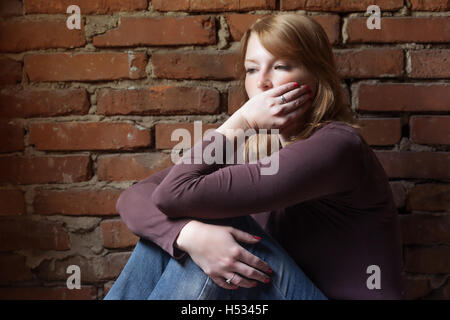 Seitlicher Blick auf eine junge rothaarige Frau, die vor einer Mauer sitzt und ist für den Mund. Stockfoto