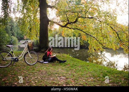 Malerischer Herbst Park. Mädchen sitzt auf dem Boden, übersät mit kleinen Herbstlaub. Herbstliche Bäume spiegeln sich in den Teich. Mädchen Stockfoto