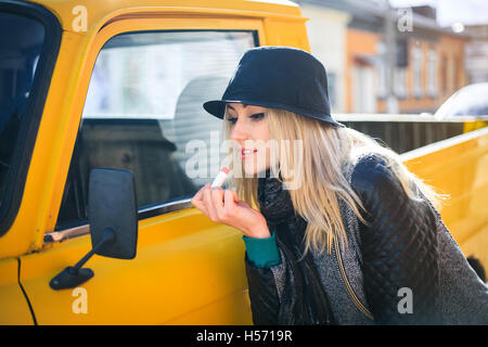 Süße junge Frau gilt roten Lippenstift betrachten die Autospiegel an einem sonnigen Herbsttag Stockfoto