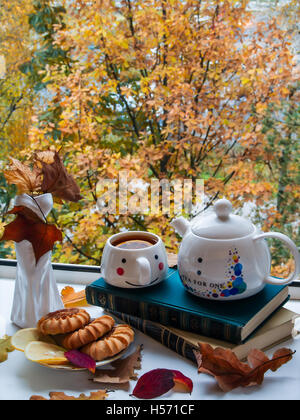 Herbst-Stilleben am offenen Fenster mit einer Vase, eine Tasse Tee, Herbstlaub, Zitrone, Bücher, Kekse Stockfoto