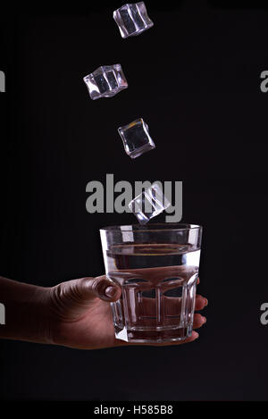 Frau hält ein Glas Wasser mit Eiswürfeln in Bewegung. Geben Sie ein Glas Wasser. Trinken Sie viel Wasser mit Eis. Schwarzer Hintergrund. Stockfoto