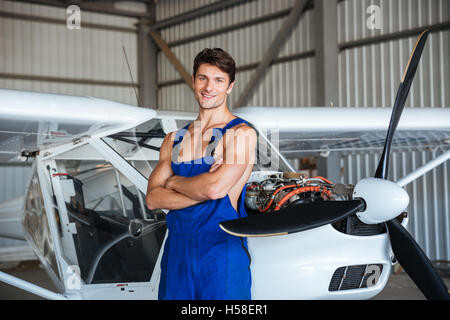 Glücklich attraktive junge Flugzeug Mechaniker stehend mit Armen überquerte in der Nähe von kleinen Flugzeug Stockfoto