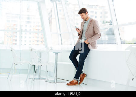 In voller Länge Portrait von lässig lächelnd Geschäftsmann stehend und mit Handy im Innenbereich Stockfoto