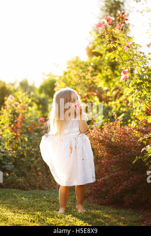 Niedliche kleine Mädchen essen eine Tomate. Sie steht barfuß auf dem grünen Rasen. Mädchen schmutzig weißes Kleid in Tomatensaft. Stockfoto