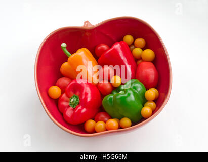 Sortierte Tomaten und rote, gelbe und grüne Paprika in eine rote Tomate geformte Schale vor einem weißen Hintergrund angeordnet Stockfoto