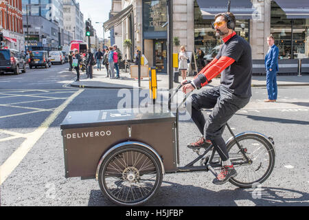 Ein Radfahrer Warenlieferungen für Deliveroo warten auf die Ampel auf Grün ändern. Ein Mann im Hintergrund auf. Stockfoto