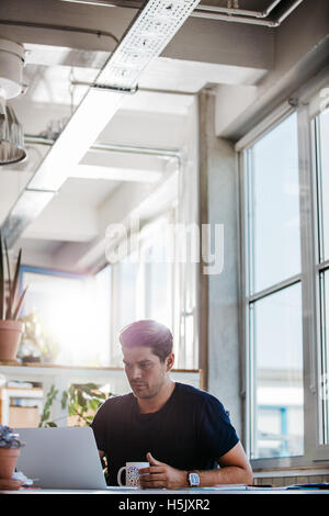 Vertikale Aufnahme von jungen kreativen Profi sitzt an seinem Schreibtisch mit Kaffee und am Laptop arbeiten. Stockfoto