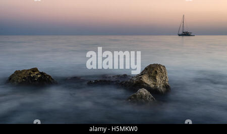 Einsame Boot am Horizont über einem felsigen Ufer im Sonnenaufgang Stockfoto