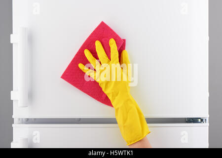 Zarte Frauenhand in schützenden gelben Latex Handschuh Reinigung weißer Kühlschrank mit rosa Lappen auf grauem Hintergrund Stockfoto