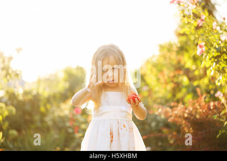 Schöne kleine Mädchen essen eine Tomate. Sie ergeht. Mädchen schmutzig weißes Kleid in Tomatensaft. Sonnenuntergang beleuchtet die wallendes Haar. Stockfoto