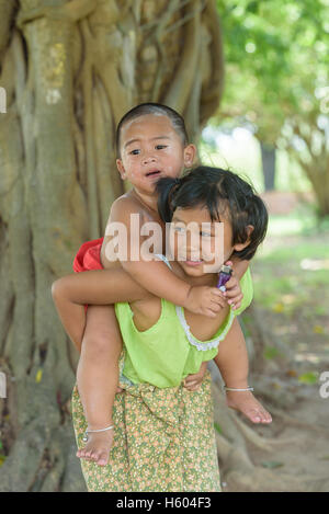 kleine asiatische junge sitzen auf junge Mädchen zurück, sie kleiden sich in Thai lokale Kleid und spielen zusammen im Freien.