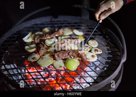 Grillen machen in der Nacht. Menschliche Hand, die Gabel, die Vermittlung von Würstchen, Kartoffeln und Zwiebeln auf Grill. Braai, Outdoor-Aktivitäten in Stockfoto