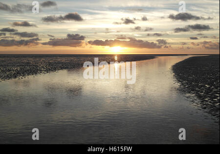 Kleine graue Stratocumulus Wolken reflektiert in einem Meer-Wasser-Kanal überqueren einer Strandsand, gegenüber einem gelben Sonnenuntergang, Fairhaven, UK Stockfoto