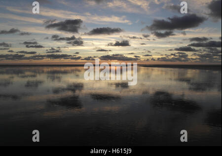 Weite Sandstrand mit vielen grauen Stratocumulus Wolken reflektiert in Meer-Wasser-Kanal in Richtung gelb Sonnenuntergang, Fairhaven, UK Stockfoto