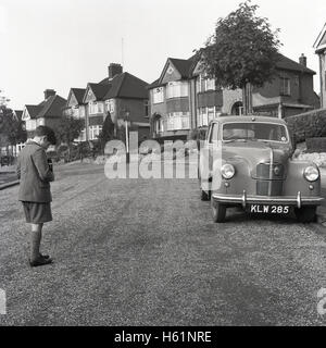 der 1950er Jahre geparkt historische, junge Schuljunge Stand in einer Straße mit dem Fotografieren in Hüfthöhe eines Kraftfahrzeugs in einer Vorstadt Straße mit einer zweiäugigen reflex Filmkamera. Stockfoto