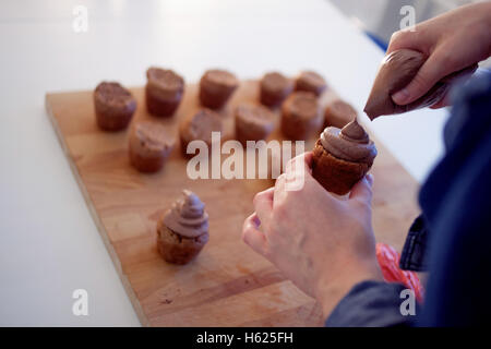 Das Mädchen deckt den Kuchen mit Sahne. Prozess des Kochens, Cupcakes, Muffins und Zutaten für die Dekoration auf dem Tisch. zarte Frauenhand hält ein cupcake Stockfoto