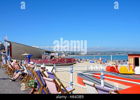 Touristen, die Erholung am Strand mit Kinder-Fahrgeschäfte und ein Café in den Vordergrund, Weymouth, Dorset, England, Vereinigtes Königreich, Western Europe. Stockfoto