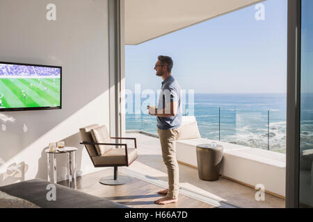 Mann beobachtet Fußball im Fernsehen an sonnigen Luxus Terrasse Tür mit Blick aufs Meer Stockfoto