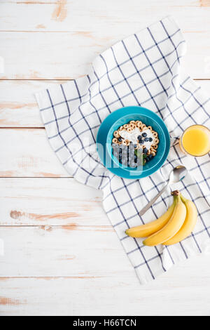 Hafer-Müsli mit Beeren und Sahne, Tasse Orangensaft, Bananen und karierte Serviette auf hölzernen Hintergrund Stockfoto