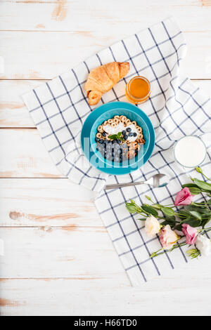 Hafer-Müsli mit Beeren und Sahne, Tasse Milch, Blumen und Plaid Serviette auf hölzernen Hintergrund Stockfoto