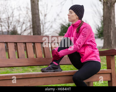 Traurige junge Sportlerin an einem kalten Wintertag in den Spuren von einem städtischen Park auf einer Bank sitzen. Stockfoto