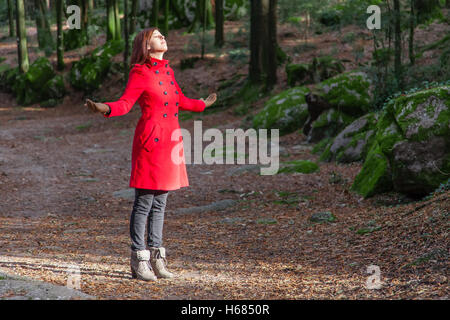 Frau mit offenen Armen genießen die Wärme im Winter Sonnenlicht an einem Wald trägt einen roten Mantel Stockfoto