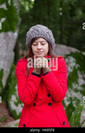 Junge Frau, die zitternd vor Kälte auf einen Wald tragen einen roten Mantel, eine Mütze und Handschuhe im winter Stockfoto