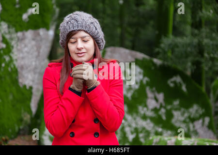 Junge Frau, die zitternd vor Kälte auf einen Wald tragen einen roten Mantel, eine Mütze und Handschuhe im winter Stockfoto