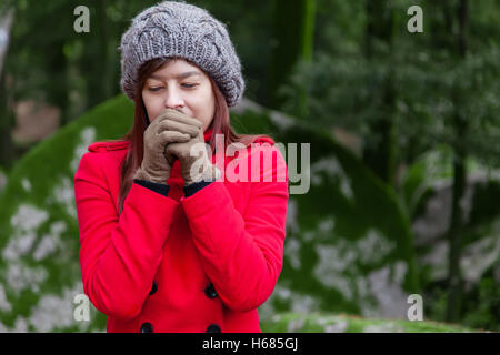 Frau, die vor Kälte zitternd und bläst warme Luft an den Händen auf einen Wald tragen einen roten Mantel, eine Mütze und Handschuhe im winter Stockfoto