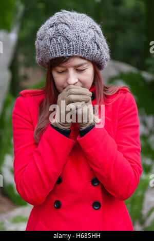 Frau, die vor Kälte zitternd und bläst warme Luft an den Händen auf einen Wald tragen einen roten Mantel, eine Mütze und Handschuhe im winter Stockfoto