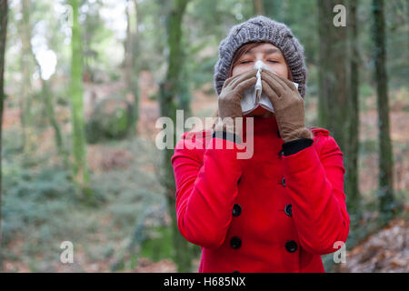 Frau mit einer Erkältung oder Grippe Niesen zu einem Papiertaschentuch auf einen Wald tragen einen roten Mantel, eine Mütze und Handschuhe im winter Stockfoto