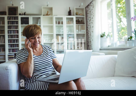 Ältere Frau am Handy während mit Laptop im Wohnzimmer Stockfoto