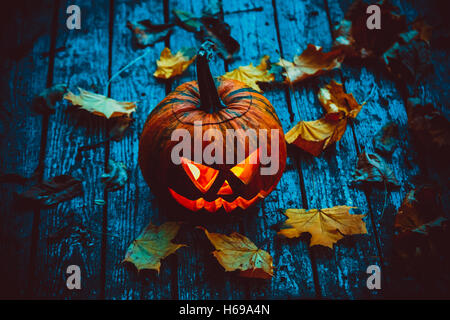 Leuchtender Kürbis, der den Kopf des alten Jack symbolisiert, mit Herbstlaub auf hölzernem Hintergrund. Weichzeichner. Oberer Freiheitsgrad Stockfoto